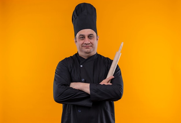 Homme d'âge moyen cuisinier en uniforme de chef tenant le rouleau à pâtisserie croisant les mains sur le mur jaune avec copie espace