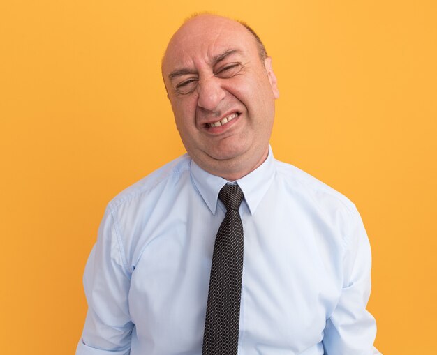 Homme d'âge moyen mécontent portant un t-shirt blanc avec une cravate isolé sur un mur orange