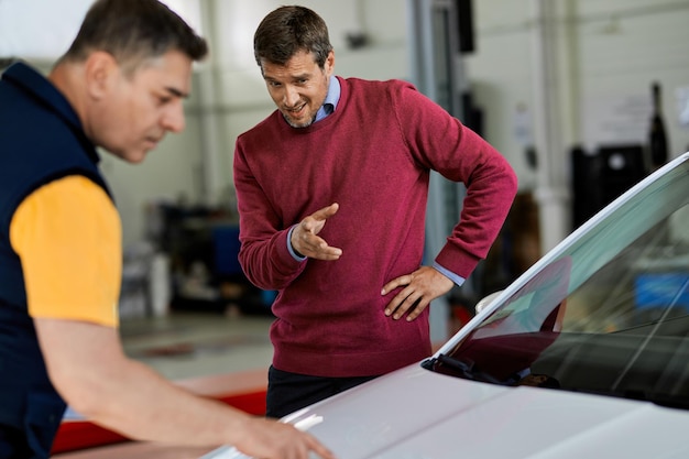 Homme d'âge moyen pointant vers le capot du véhicule tout en parlant à un mécanicien automobile dans un atelier de réparation automobile