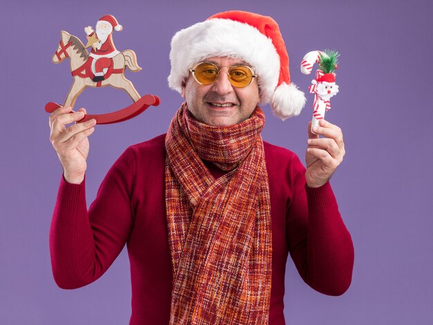 Homme d'âge moyen portant bonnet de noel de Noël avec écharpe chaude autour du cou dans des verres jaunes montrant des jouets de Noël heureux et joyeux souriant debout sur fond violet