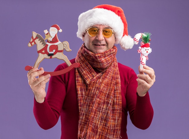 Homme d'âge moyen portant bonnet de noel de Noël avec écharpe chaude autour du cou dans des verres jaunes montrant des jouets de Noël heureux et joyeux souriant debout sur fond violet