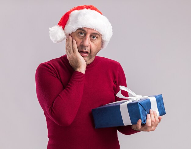 Homme d'âge moyen portant un bonnet de noel de noël tenant un cadeau de noël surpris et étonné debout sur un mur blanc