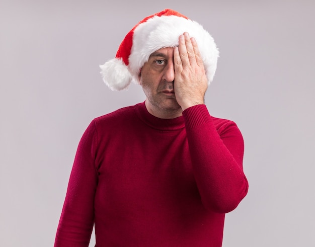 Homme d'âge moyen portant un bonnet de noel avec un visage sérieux couvrant un vous avec la main debout sur un mur blanc