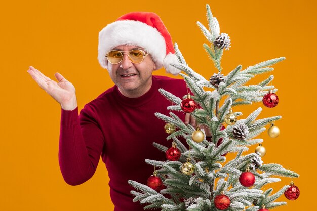 Homme d'âge moyen portant chapeau de Père Noël en col roulé rouge foncé et lunettes jaunes décoration arbre de Noël regardant la caméra souriant joyeusement debout sur fond orange
