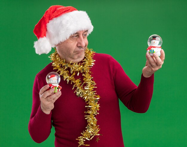 Homme d'âge moyen portant chapeau de Père Noël avec guirlandes autour du cou tenant des boules de neige de Noël à la confusion essayant de faire un choix