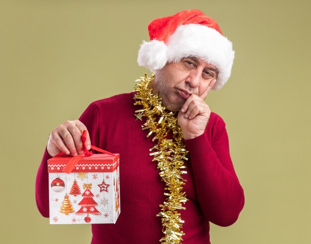 Homme d'âge moyen portant chapeau de père Noël avec guirlandes autour du cou tenant le cadeau de Noël regardant la caméra avec la main sur le menton pensant debout sur fond vert