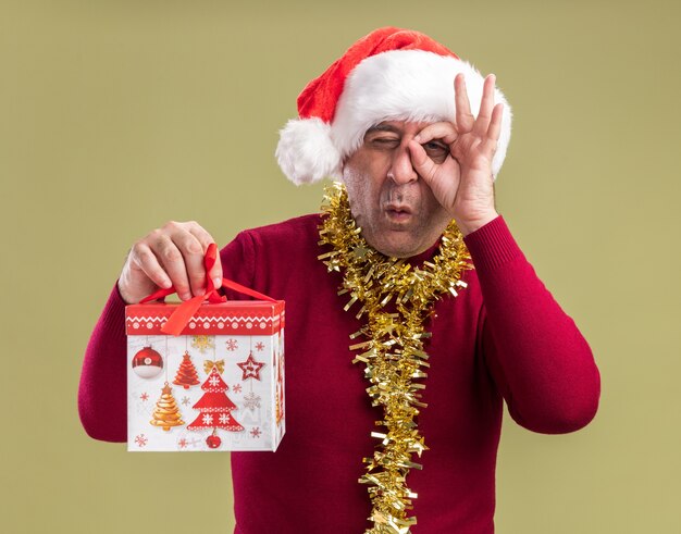 Homme d'âge moyen portant chapeau de Père Noël avec guirlandes autour du cou tenant un cadeau de Noël regardant la caméra à travers les doigts faisant ok chanter debout sur fond vert