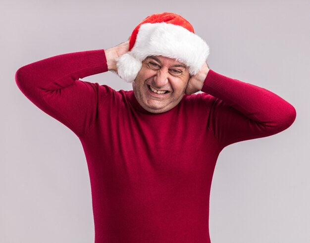 Homme d'âge moyen portant noël santa hat fermant les oreilles avec les mains avec une expression agacée debout sur fond blanc