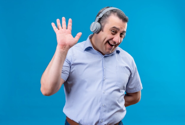 Homme d'âge moyen positif et joyeux en chemise rayée bleue portant des écouteurs montrant cinq gestes sur un fond bleu
