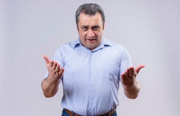 Homme d'âge moyen sérieux en chemise à rayures verticales bleu surpris et posant des questions avec la main posée sur un fond blanc