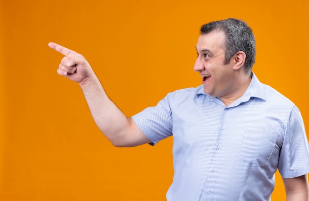 Homme d'âge moyen souriant et positif en chemise à rayures verticales bleu pointant vers le coin supérieur droit avec l'index en se tenant debout sur un fond orange