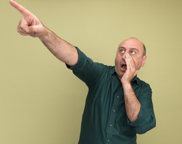 Un homme d'âge moyen surpris portant des points de t-shirt vert sur le côté appelant quelqu'un isolé sur un mur vert olive