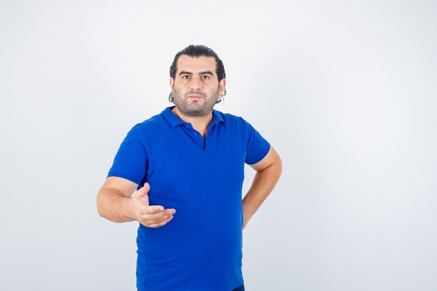 Homme d'âge moyen en t-shirt bleu qui s'étend de la main en geste de questionnement et à la vue de face, confiant.