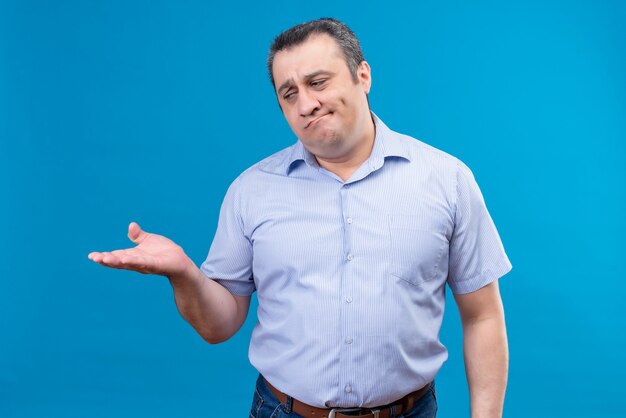 Homme d'âge moyen troublé et bouleversé en chemise rayée bleue levant la main sur un fond bleu