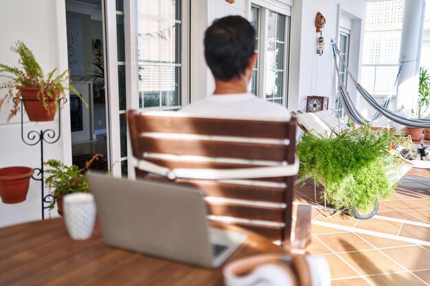 Homme d'âge moyen utilisant un ordinateur portable à la maison debout en arrière regardant loin avec les bras croisés