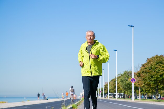 Homme d'âge mûr ciblé en veste de sport verte et collants jogging le long de la rivière à l'extérieur. Formation de jogger senior pour marathon. Vue de face. Concept d'activité et d'âge