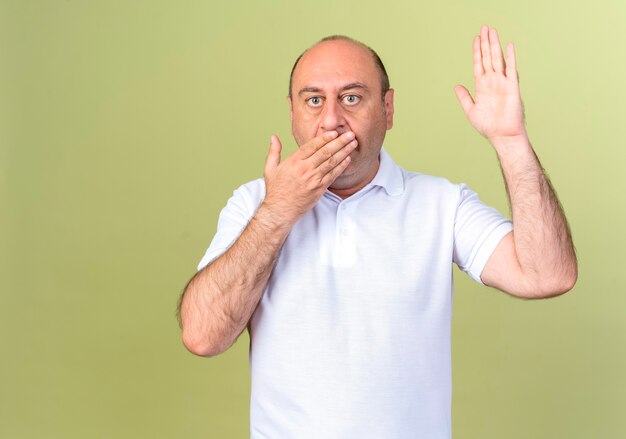 Homme d'âge mûr effrayé bouche couverte et main levée isolé sur mur vert olive