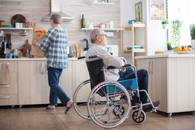 Un homme âgé non valide souriant en regardant par la fenêtre dans la cuisine et sa femme déballe les courses. Invalide, retraité, handicapé, paralysie.