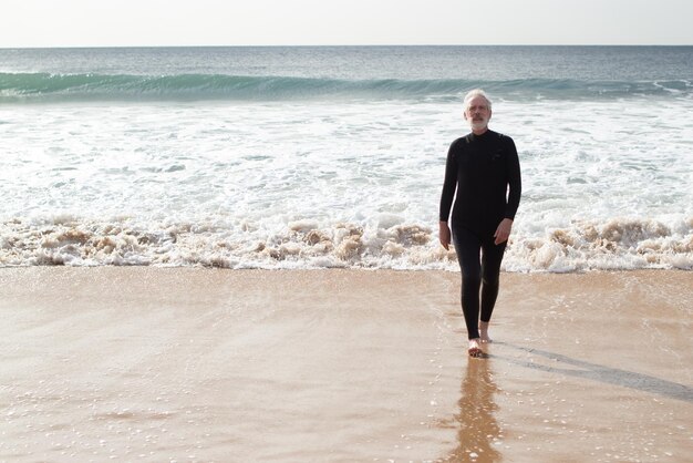 Homme âgé sérieux ayant un repos actif près de la mer. Homme sportif aux cheveux gris en combinaison de plongée sortant de la mer après avoir nagé en marchant pieds nus en regardant la caméra. Sport et mode de vie sain du concept de personnes âgées