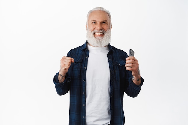 Photo gratuite un homme âgé souriant dit oui, fait une pompe à poing, serre les poings en triomphe et tient un smartphone, rit en gagnant et célèbre le succès, atteint l'objectif en ligne dans l'application, mur blanc