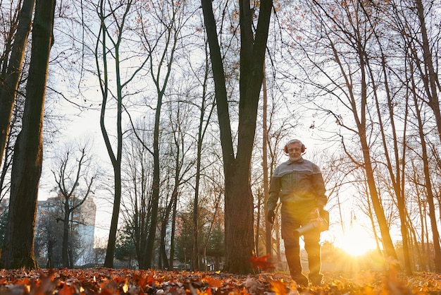 Photo gratuite homme âgé souriant travaillant avec un souffleur de feuilles lourd, les feuilles brillent sous un soleil agréable, vue à angle bas