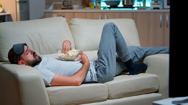 Homme allongé dans le canapé en train de manger du popcron et de regarder la télévision