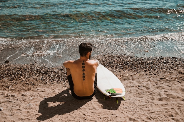 Homme anonyme avec planche de surf assis sur une plage de sable