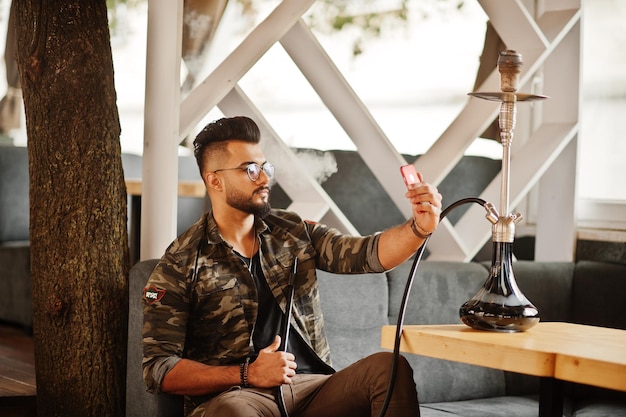 Homme arabe barbe élégante dans des verres et une veste militaire fumant le narguilé au bar de la rue Modèle arabe se reposant et prenant un selfie au téléphone