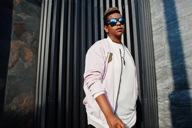 Homme arabe hipster élégant à lunettes de soleil posé en plein air dans la rue au coucher du soleil contre un mur gris