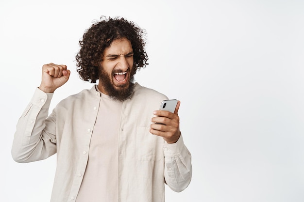 Un homme arabe joyeux regardant un téléphone portable et célébrant le fait de regarder une vidéo de jeu de sport sur un smartphone debout sur fond blanc