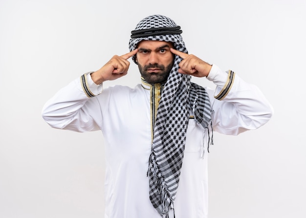 Homme arabe en tenue traditionnelle pointant ses tempes avec une expression confiante sérieuse debout sur un mur blanc
