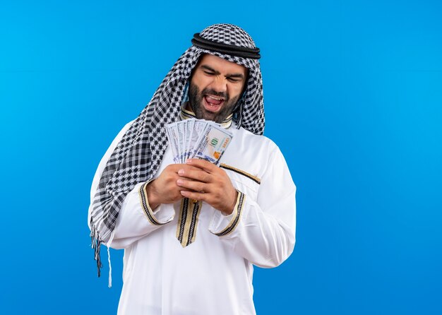 Homme arabe en tenue traditionnelle tenant de l'argent en souriant joyeusement debout sur un mur bleu