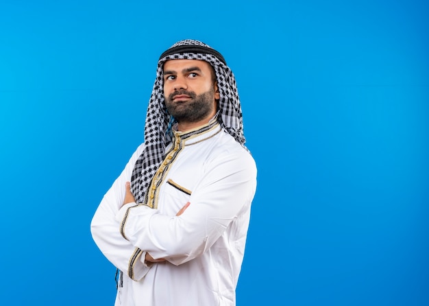 Photo gratuite l'homme arabe en vêtements traditionnels à côté avec une expression confiante avec les mains croisées sur la poitrine debout sur le mur bleu