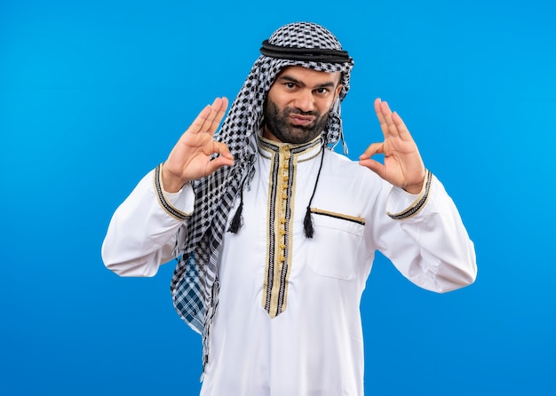 L'homme arabe en vêtements traditionnels faisant des signes oo avec les deux mains à la confiance debout sur le mur bleu