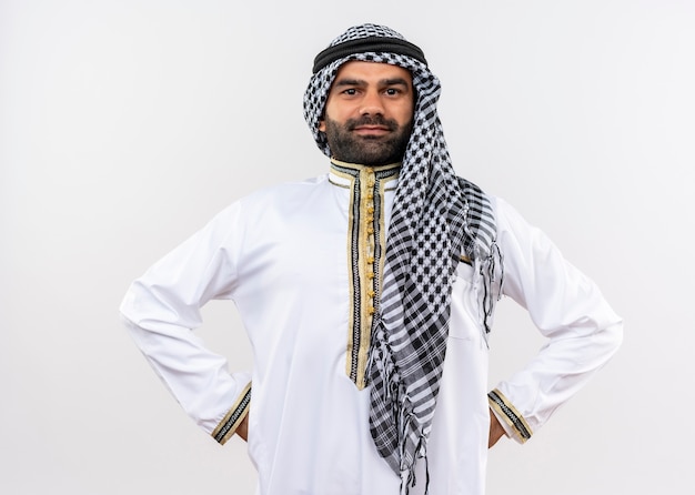 Homme arabe en vêtements traditionnels avec un sourire confiant sur le visage debout sur un mur blanc