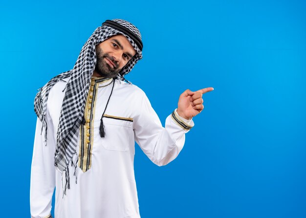 L'homme arabe en vêtements traditionnels avec sourire sur le visage pointant avec le doigt sur le côté debout sur le mur bleu