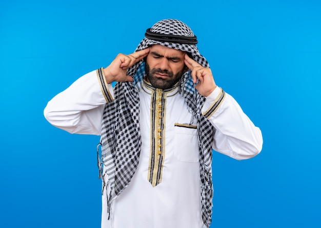 L'homme arabe en vêtements traditionnels touchant ses tempes souffrant de forts maux de tête debout sur un mur bleu