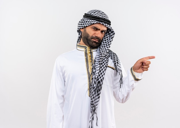 Homme arabe en vêtements traditionnels avec visage fronçant pointant avec le doigt sur le côté debout sur un mur blanc