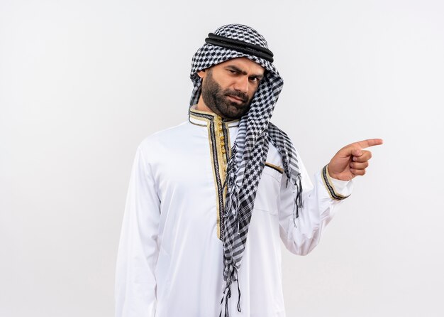 Homme arabe en vêtements traditionnels avec visage fronçant pointant avec le doigt sur le côté debout sur un mur blanc