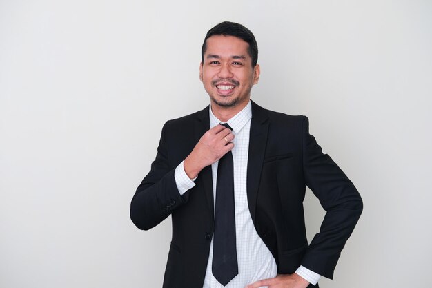 Homme asiatique adulte portant un costume noir montrant une grande confiance avec la main touchant sa cravate