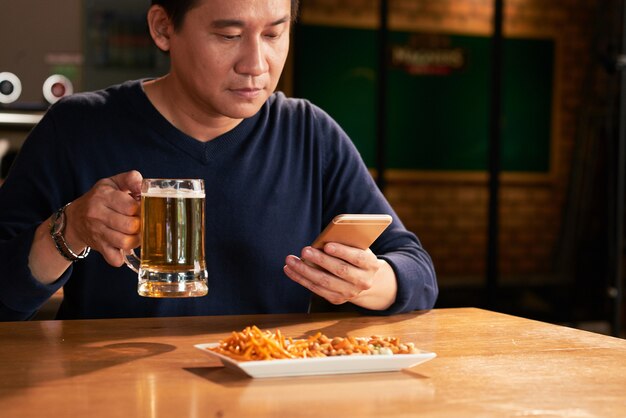 Homme asiatique assis dans un pub avec de la bière et des collations et à l'aide de smartphone