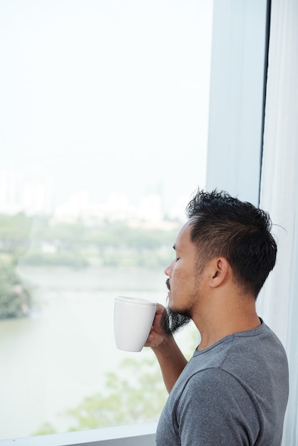 Homme asiatique, debout, devant, fenêtre, et, sentant, tasse café, à, yeux fermés