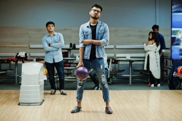Homme asiatique élégant en veste de jeans et lunettes debout au bowling avec ballon à portée de main