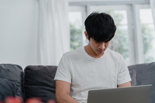 Homme asiatique indépendant travaillant à la maison, créatif masculin sur un ordinateur portable sur le canapé dans le salon. Entrepreneur entrepreneur jeune homme d'affaires, jouer à l'ordinateur, vérifier les médias sociaux en milieu de travail à la maison moderne.