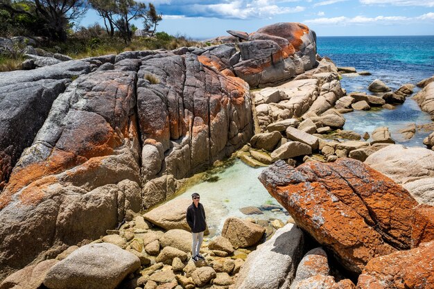 Un homme asiatique pose pour la caméra en se tenant debout sur de gros rochers à côté d'une mer