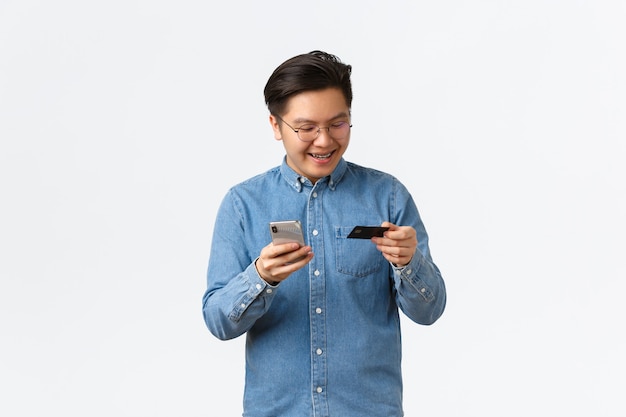Homme asiatique souriant et insouciant dans des lunettes et des vêtements décontractés, ayant des bretelles, regardant une carte de crédit avec plaisir, achetant en ligne avec une application pour smartphone, faisant des achats sur Internet avec un téléphone portable.