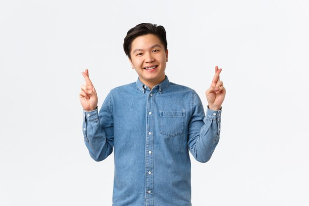 Un homme asiatique souriant et mignon ayant la foi, croit que les rêves deviennent réalité, croise les doigts bonne chance, prie ou fait un vœu, se tient chanceux sur fond blanc, anticipe des résultats positifs.