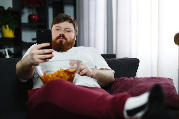 Photo gratuite l'homme assez gros sourit en vérifiant son smartphone alors qu'il est assis sur le canapé et mange