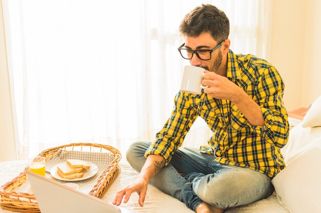 Photo gratuite homme assis sur un lit, buvant le café avec petit-déjeuner et ordinateur portable sur le lit