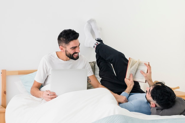 Homme assis avec un ordinateur portable près de son petit ami en lisant le livre sur le lit